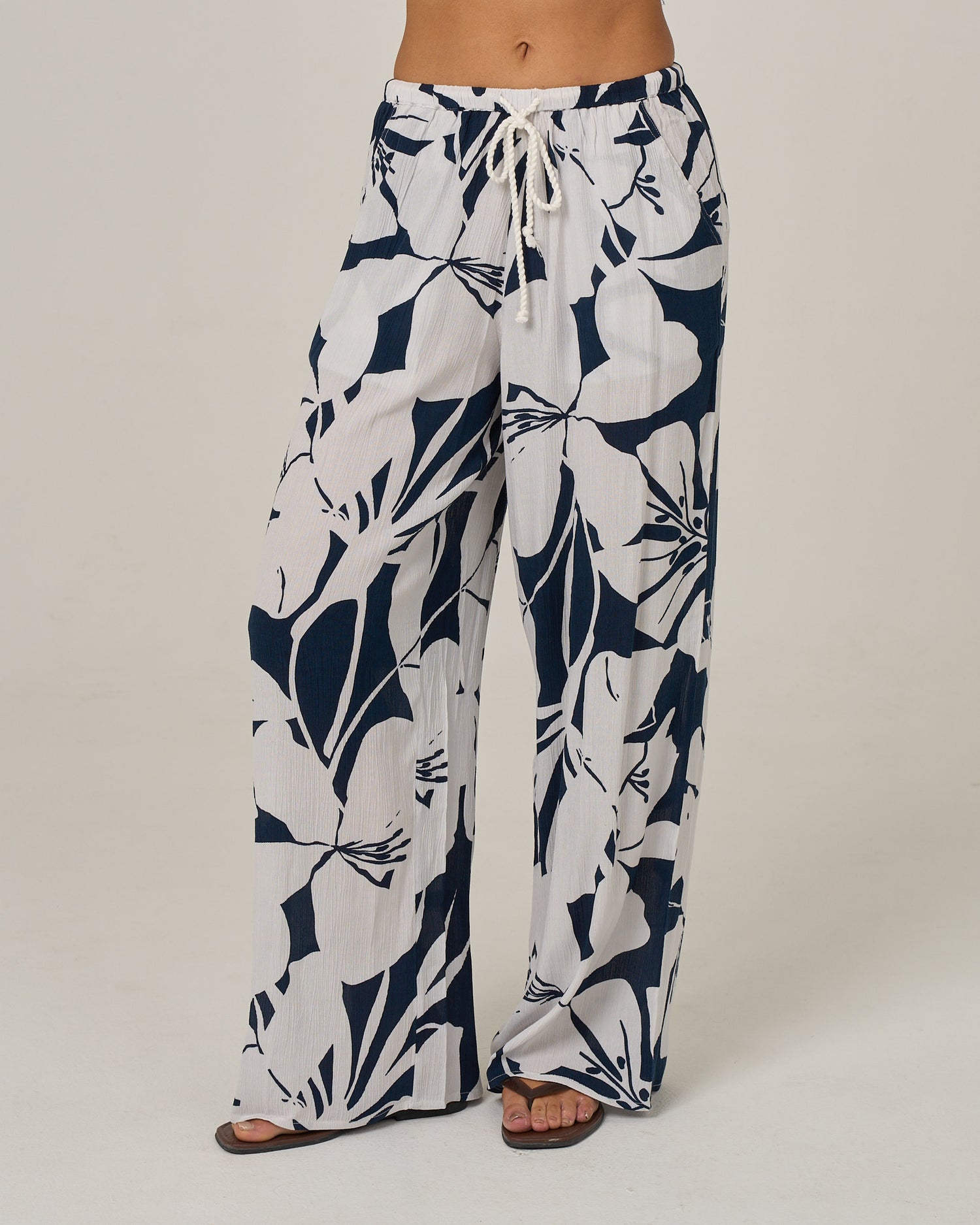 A person wearing the Salty Crew Graphic Bloom Pant in Dark Navy, featuring a loose, wide-leg silhouette with a navy and white tropical leaf print, an elastic waistband with a white rope tie drawcord, paired with open-toe sandals.
