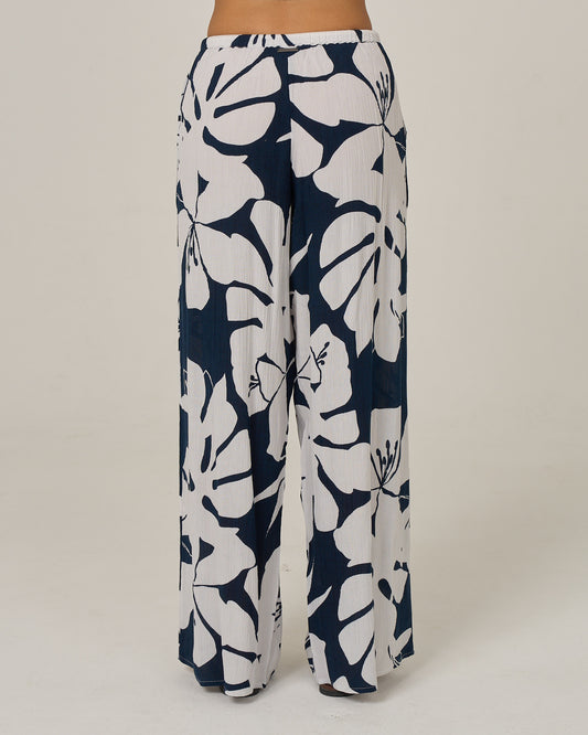 A person is shown from the back against a plain light background, wearing the Salty Crew Graphic Bloom Pant in Dark Navy—wide-leg pants with a bold navy blue and white floral print, featuring an elastic waist and rope tie drawcord.