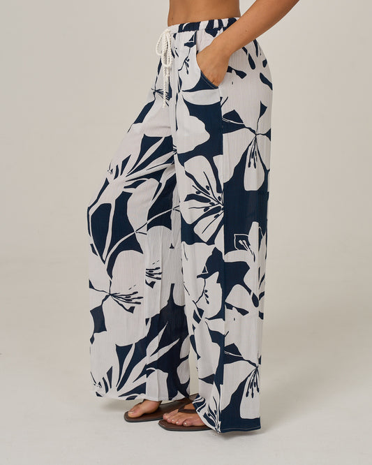 A person wearing the Salty Crew Graphic Bloom Pant in Dark Navy, featuring white wide-leg viscose fabric with large navy floral prints, pockets, and an elastic waist with a rope tie drawcord, paired with black sandals and standing against a plain light background.