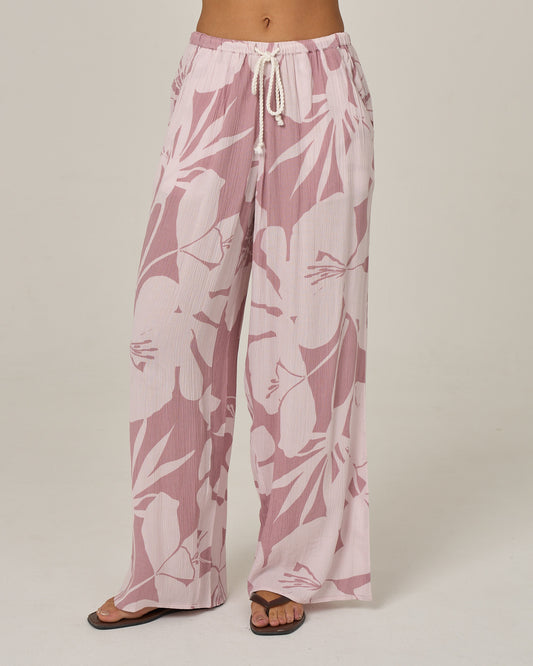 A woman stands on a plain background wearing Salty Crew’s Graphic Bloom Pant in Faded Mauve, a loose-fitting viscose beach pant featuring a light pink and white floral print, elastic waistband, trouser pockets, and paired with black sandals.