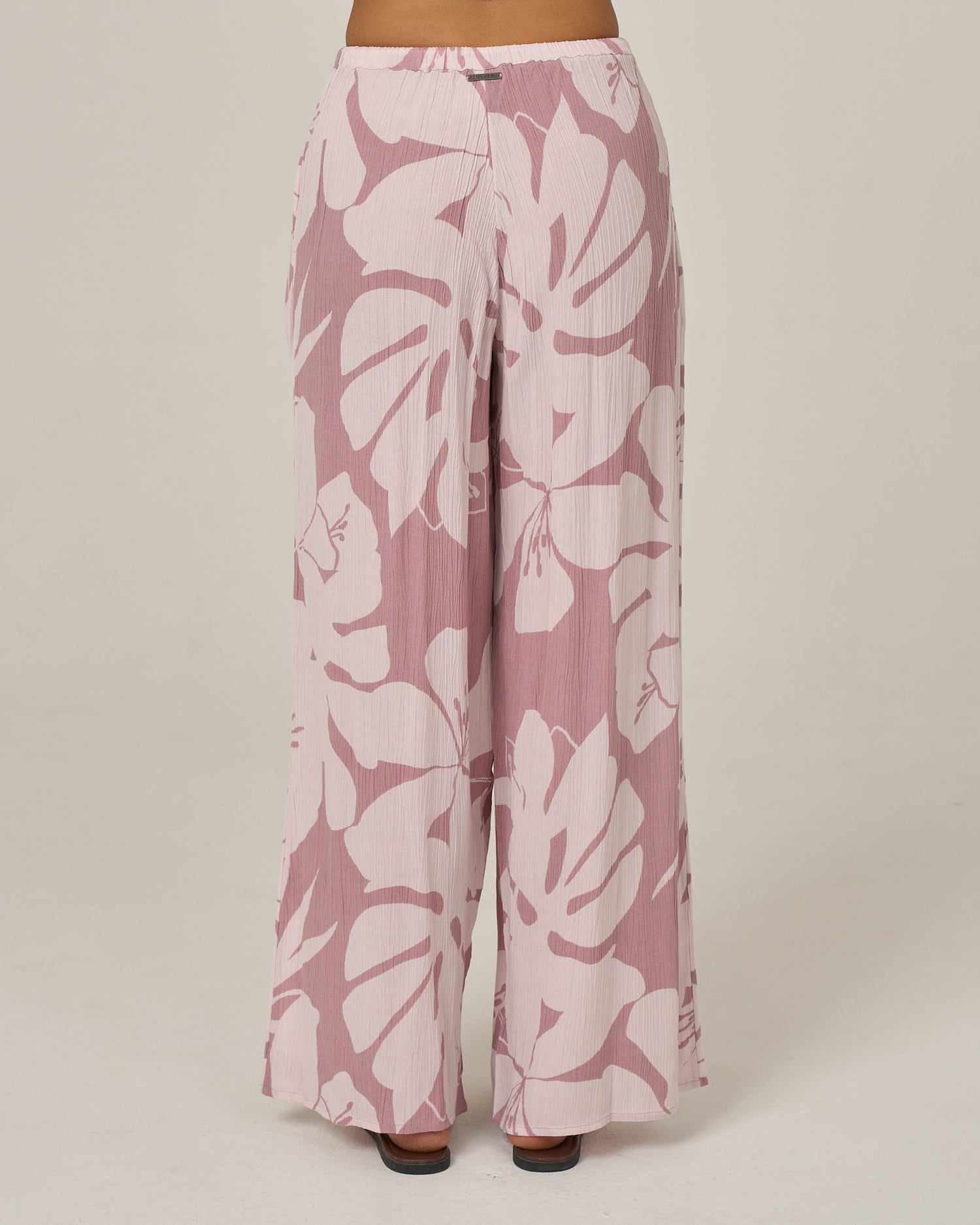 The Graphic Bloom Pant - Faded Mauve by Salty Crew is a wide-leg viscose beach pant with a light pink and white tropical leaf print, featuring an elastic waist, worn by a person standing against a plain off-white background.