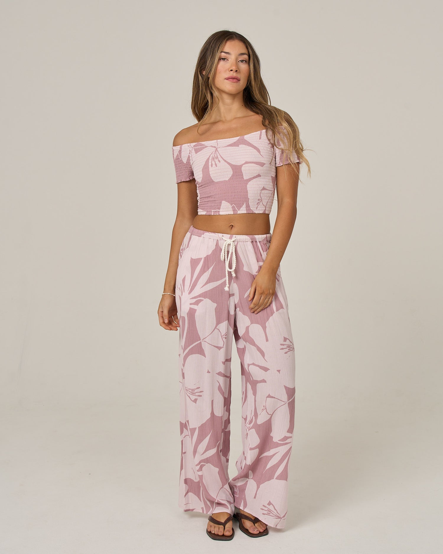 A woman stands against a plain background wearing the Salty Crew Graphic Bloom Pant in Faded Mauve, featuring a relaxed wide-leg silhouette, elastic waist, and practical pockets. She pairs the pants with a pink and white off-the-shoulder crop top. Her hair is loose and she wears black sandals, finishing the laid-back look with a neutral expression.