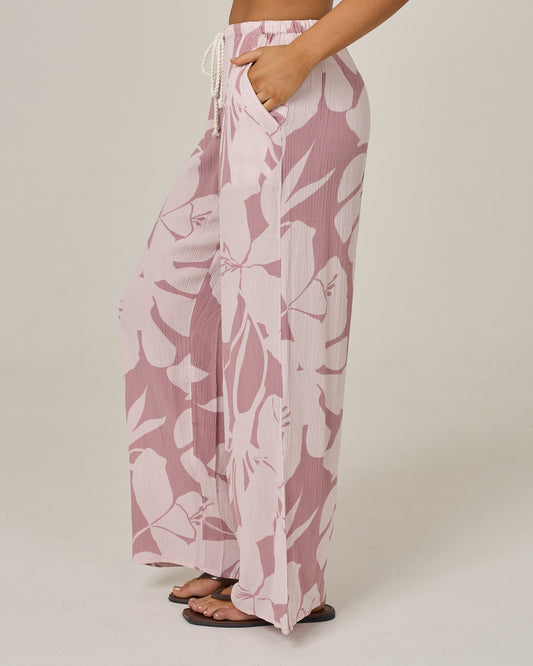 A person is shown standing sideways against a plain background, wearing the Salty Crew Graphic Bloom Pant in Faded Mauve—loose, wide-leg floral print viscose beach pants featuring an elastic waist and trouser pockets—paired with flat sandals.