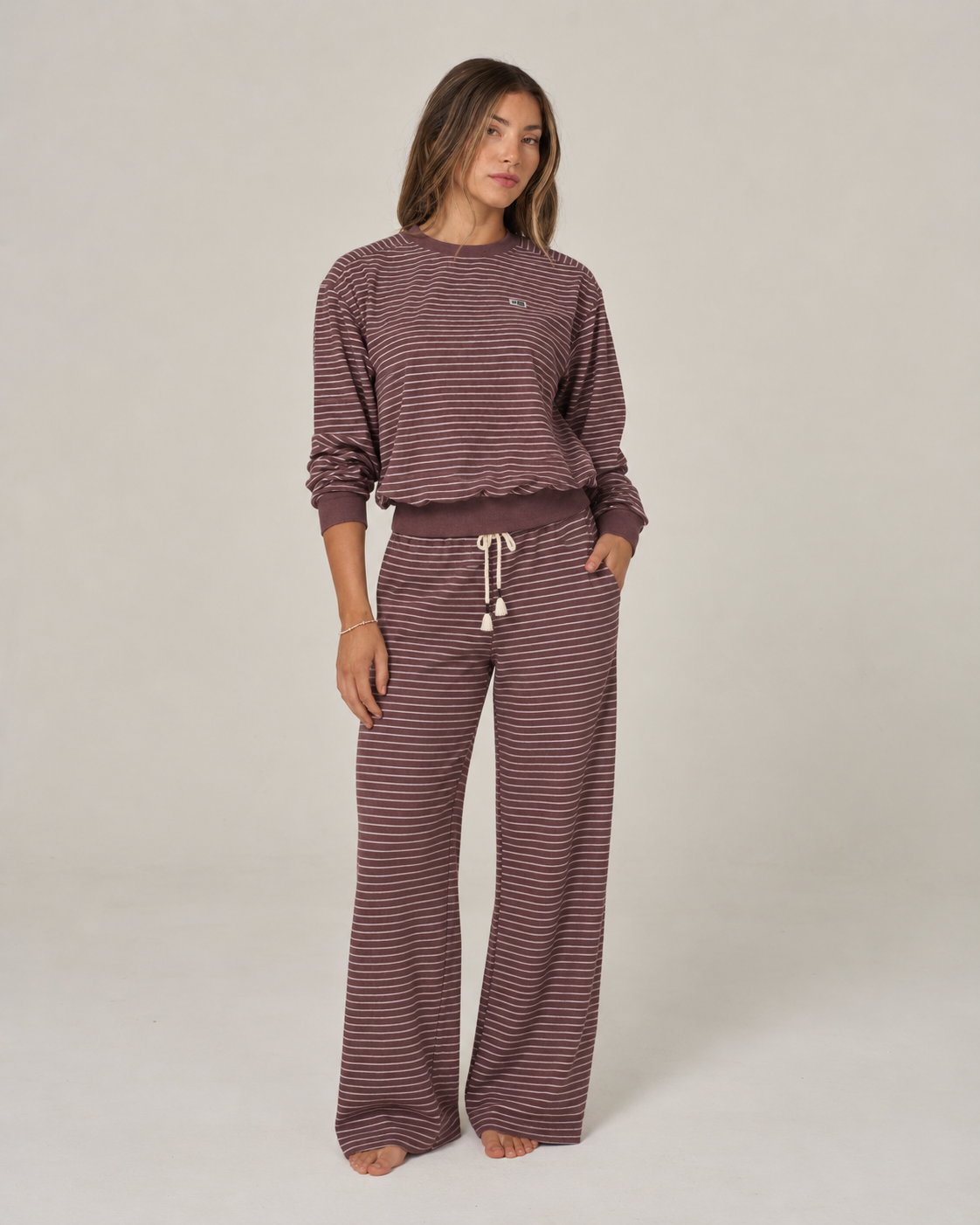 A woman stands barefoot on a plain background, looking slightly to the side with a relaxed expression. She is wearing the Salty Crew Leeward Pant in maroon—a wide-leg terry stripe pant with an elastic waist—paired with a matching long-sleeved, loose-fitting brown and pink striped top.