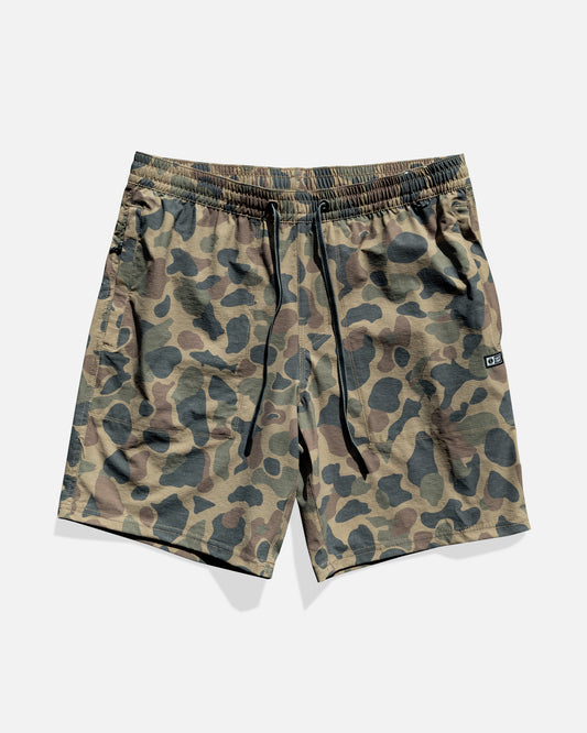The Salty Crew Crew 18" Boatshort Ewaist in Sand Camo features green and brown camo, elastic waistband with black drawstrings, a logo patch on the left leg, and is made from quick-dry ripstop fabric. Shorts are shown flat on a white background.