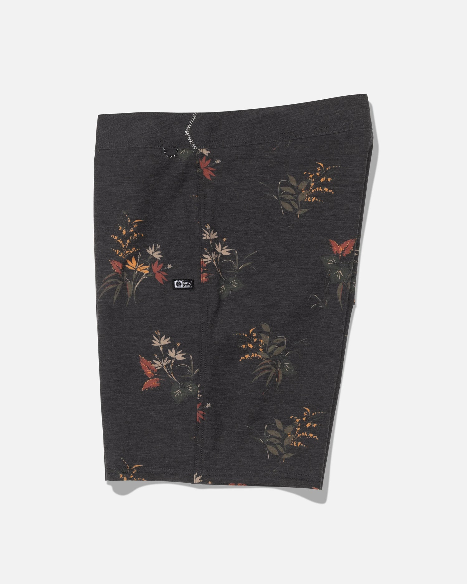 The Crew 19″ Boardshort - Off Black by Salty Crew is crafted from recycled polyester with 4-way stretch for optimal movement. These dark gray boardshorts feature a subtle floral pattern with red, orange, and yellow flowers, along with green leaves. Displayed flat against a white background, they include a small black logo patch on the left side.