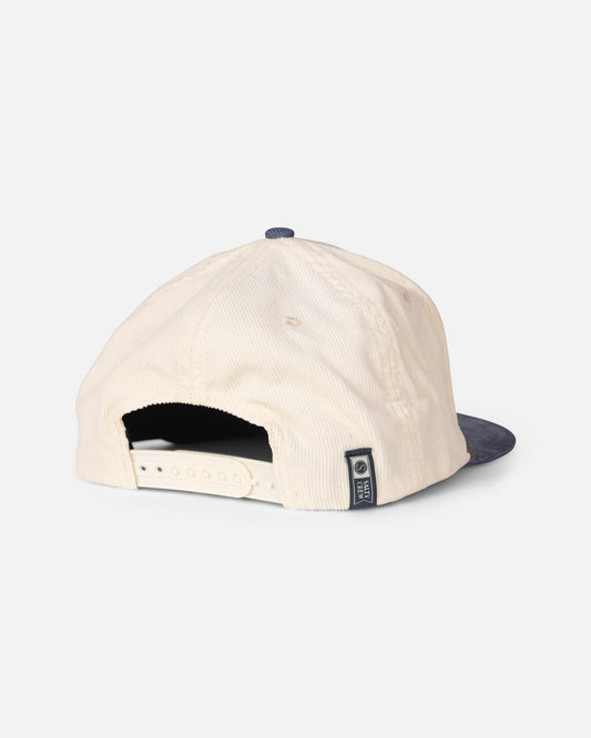The Salty Crew Tarpon Snapback - Off White Blue features a classic baseball cap style in cream-colored cotton corduroy, paired with a navy blue brim. Viewed from the back, it showcases an adjustable snapback strap and a small stitched label on the side.