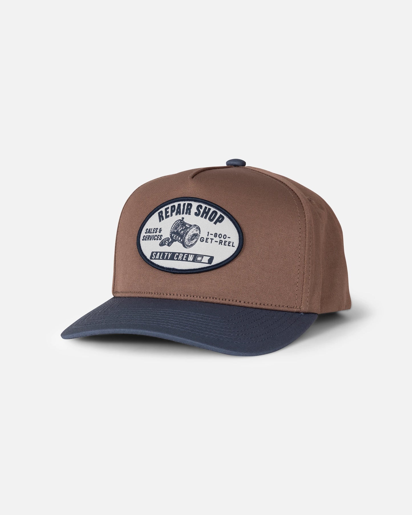 Introducing the Salty Crew Reel Repair Snapback in Brown/Navy—a structured 5-panel baseball cap crafted from durable cotton canvas. This snapback features a patch on the front reading “Repair Shop, Sales Service, 1-800-GET-REEL, Salty Crew,” along with an image of a reel. Photographed at an angle against a white background.