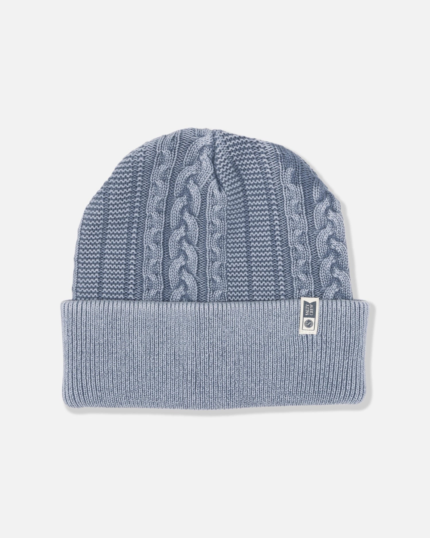 The Salty Crew Fisherman Beanie - Flint is a light blue cable knit beanie featuring a folded brim and a vertical cable knit pattern. Crafted in a classic cotton style, it includes a small white rectangular tag with dark text sewn onto the brim. The beanie is shown against a plain white background.