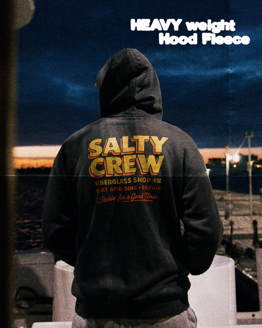 A person in a dark hoodie with Salty Crew text on the back stands on a boat at sunset. The sky glows blue and orange. White text reads: Fiberglass Heavyweight Fleece - Washed Black—crafted from premium brushed terry fleece.