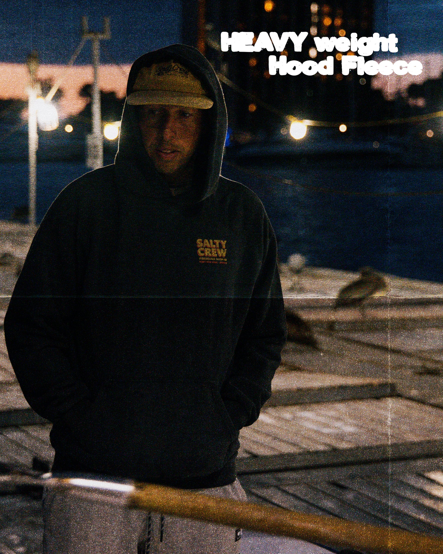 A man in a Salty Crew Fiberglass Heavyweight Fleece in washed black and a tan cap stands on a dock at dusk, with water and boats behind him. In the upper right, text reads: HEAVY weight Hood Fleece.