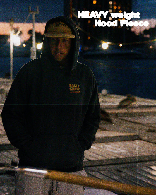 A man in a Salty Crew Fiberglass Heavyweight Fleece in washed black and a tan cap stands on a dock at dusk, with water and boats behind him. In the upper right, text reads: HEAVY weight Hood Fleece.