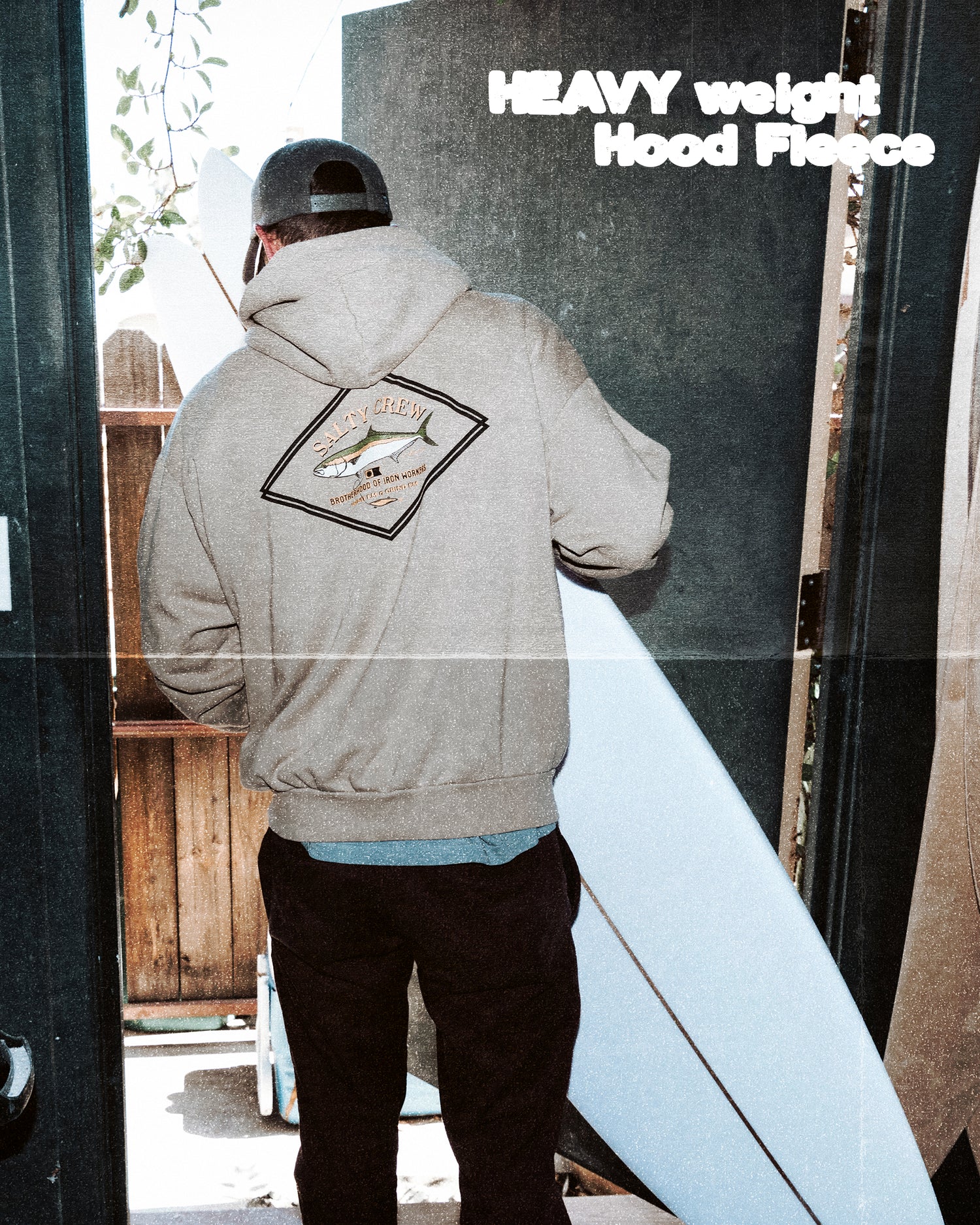 A person wearing the Salty Crew Yellowtail Heavyweight Fleece in Washed Stone, featuring a back graphic, stands in a doorway holding a white surfboard. “HEAVY weight Hood Fleece” appears in the top right corner.