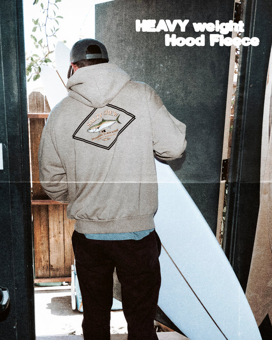 A person wearing the Salty Crew Yellowtail Heavyweight Fleece in Washed Stone, featuring a back graphic, stands in a doorway holding a white surfboard. “HEAVY weight Hood Fleece” appears in the top right corner.