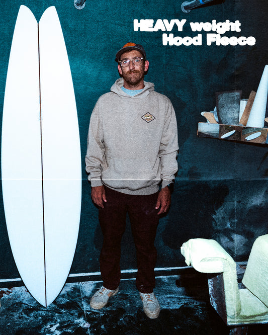 A man in glasses and a hoodie stands in a workshop by a white surfboard blank, tools, and boards. Text reads Salty Crew Yellowtail Heavyweight Fleece - Washed Stone, crafted from 100% cotton fleece, at the top right.