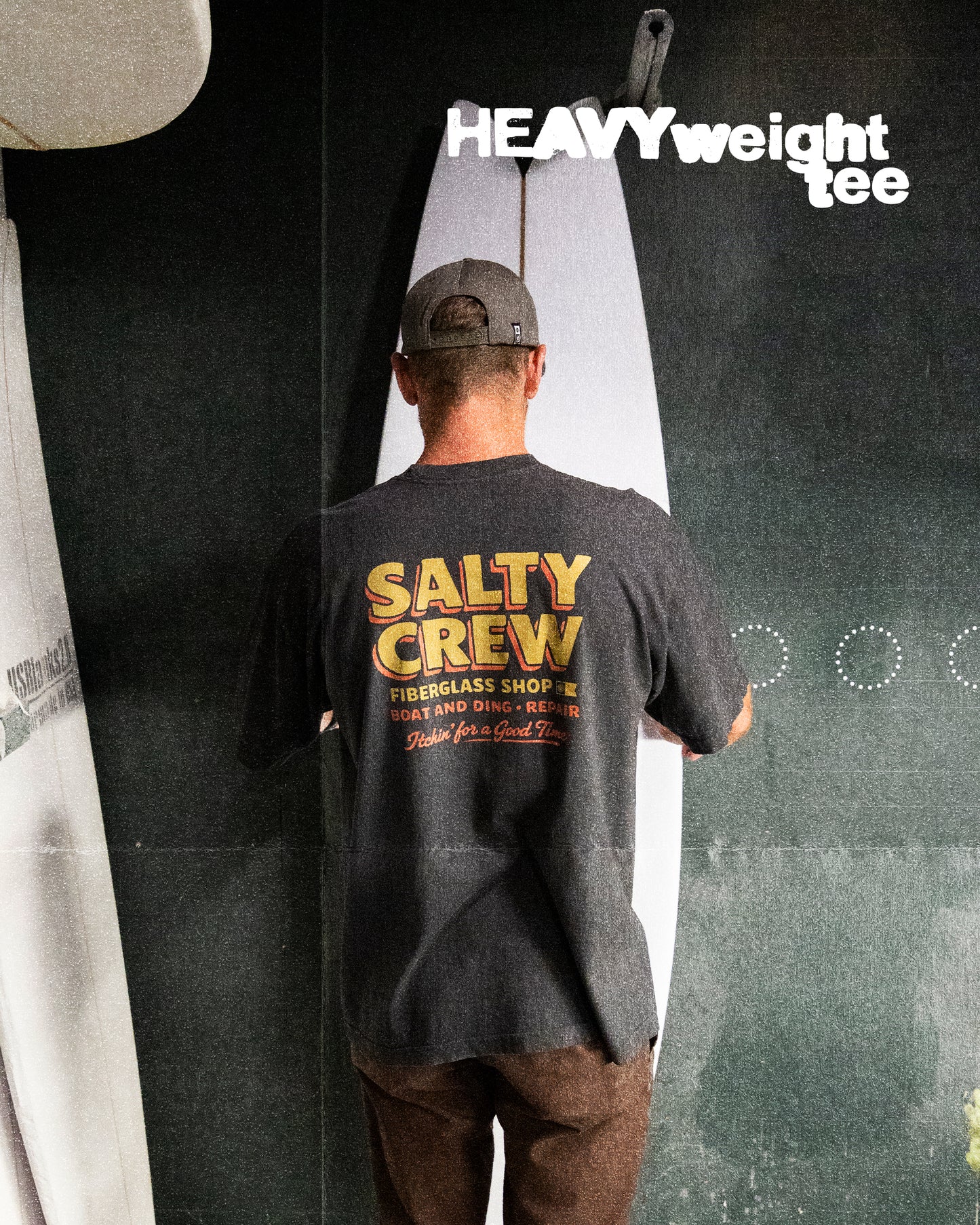 A man faces away from the camera, wearing a Salty Crew Fiberglass Heavyweight Tee in washed black, made from 100% cotton, standing in front of a surfboard. “HEAVYweight tee” is displayed in bold white at the top right.