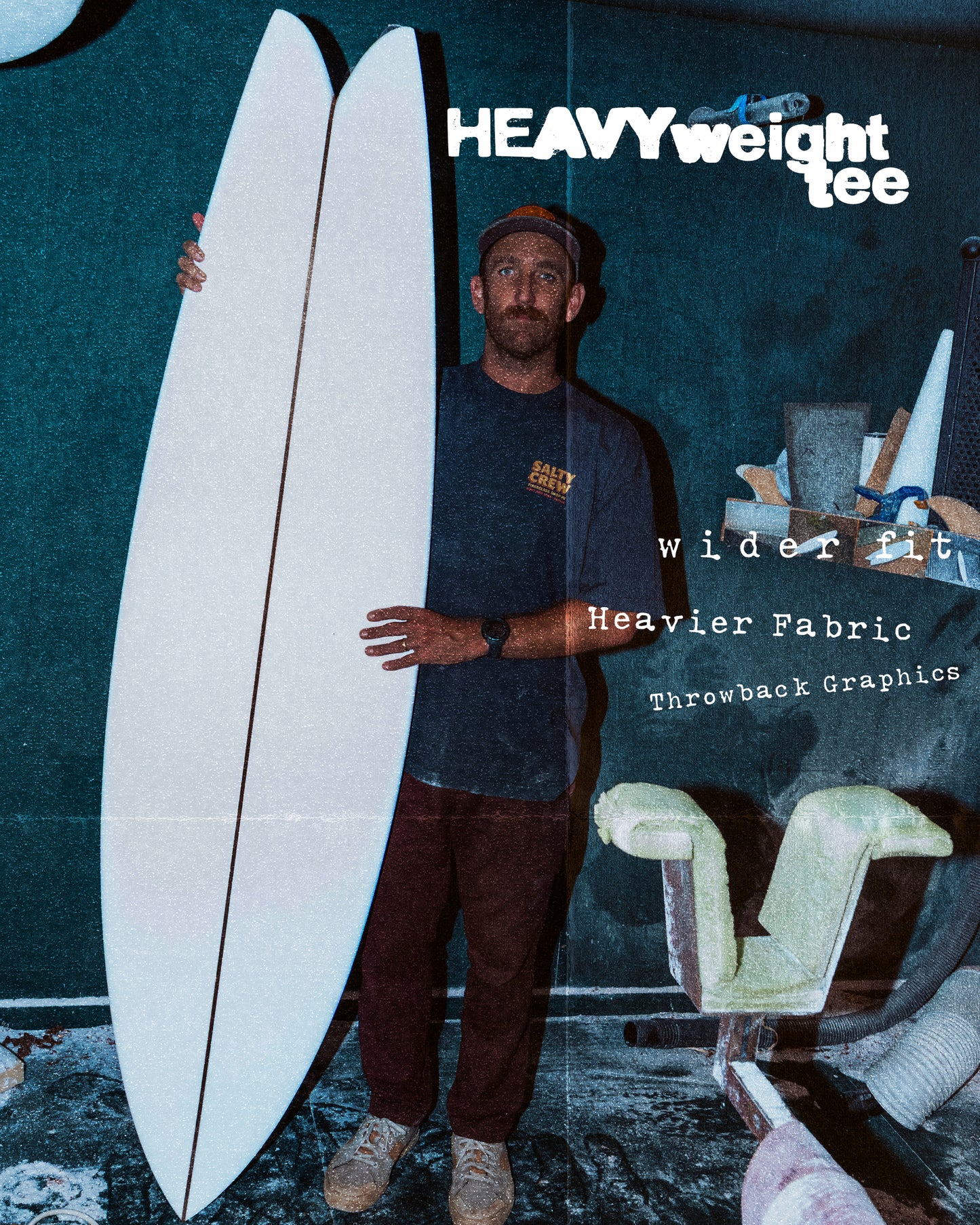 A man stands indoors holding a large, white surfboard. Bold text reads Fiberglass Heavyweight Tee - Washed Black by Salty Crew, featuring screenprinted throwback graphics, a wider fit, and heavier 100% cotton for lasting comfort.