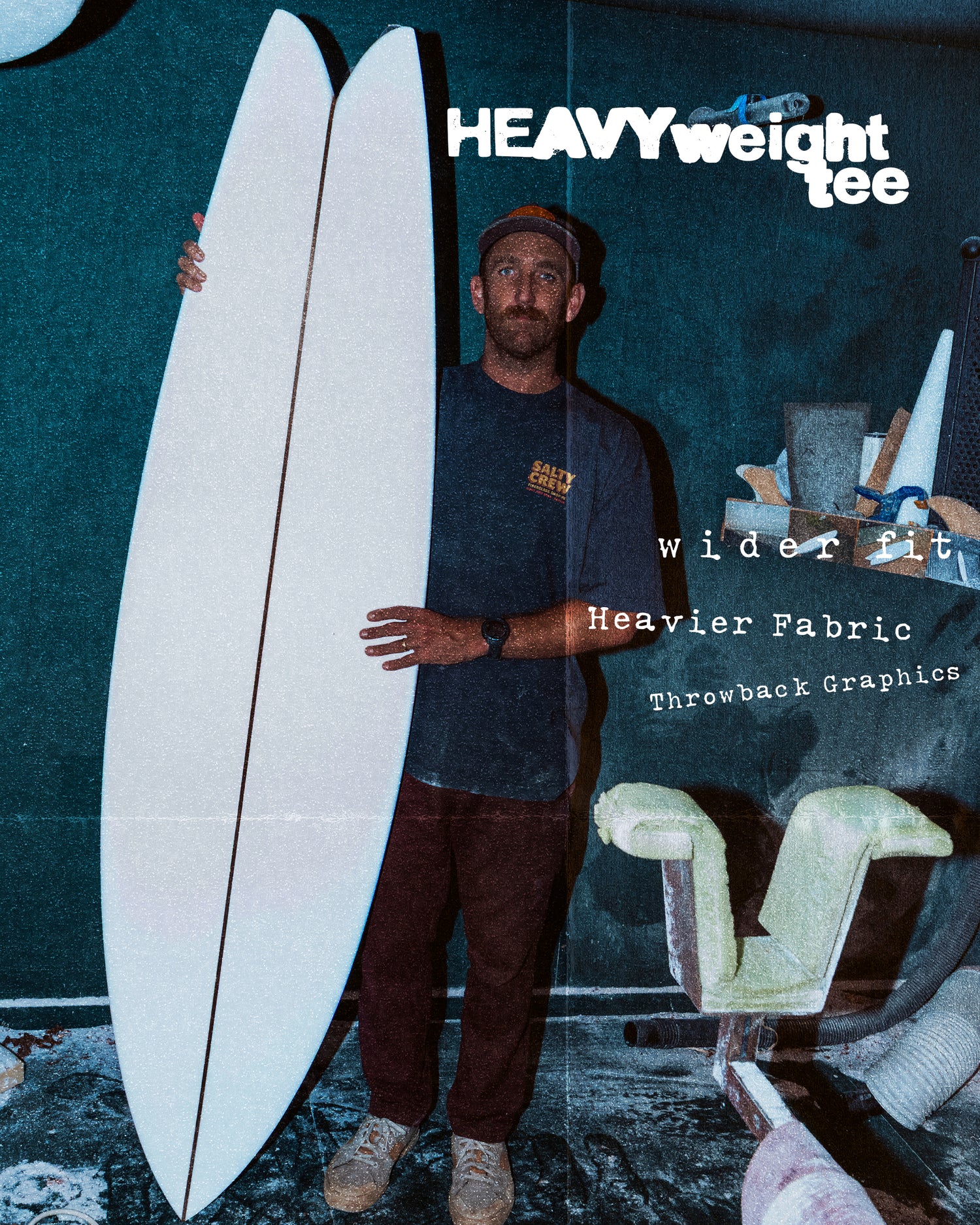 A man stands indoors holding a large, white surfboard. Bold text reads Fiberglass Heavyweight Tee - Washed Black by Salty Crew, featuring screenprinted throwback graphics, a wider fit, and heavier 100% cotton for lasting comfort.