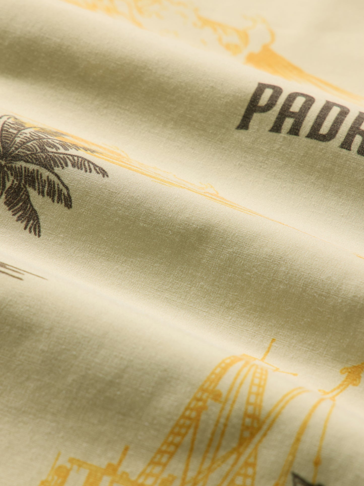 A close-up view of the Padres x Reyn Spooner UV Button Up in Bone by Salty Crew highlights its cream-colored fabric adorned with yellow and brown illustrations, such as a palm tree, a bridge, and partial PADR text. The softly wrinkled surface embodies the laid-back coastal style that Salty Crew is known for—making it a perfect choice for San Diego Padres fans looking to channel relaxed West Coast vibes.