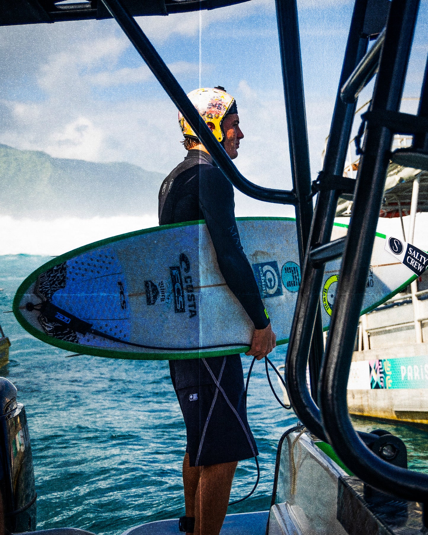 A surfer in a helmet stands on a boat, holding a surfboard and wearing Salty Crew’s Portal 21" Boardshort - Black, gazing at the ocean with waves and mountains behind. Surfboards and boat gear can be seen nearby.