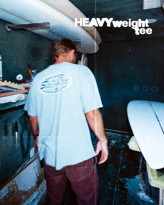A man stands in a surfboard workshop wearing the Salty Crew Service Heavyweight Tee in washed steel grey, featuring a large back graphic and bold white ‘HEAVYweight Tee’ text on the top right. Unfinished boards surround him.