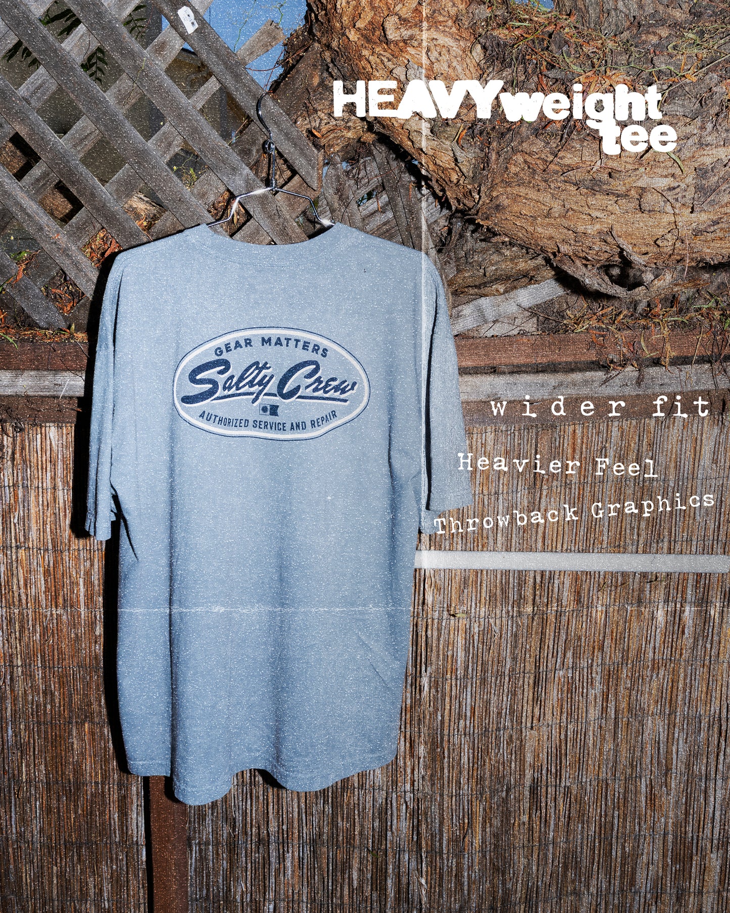 A Salty Crew Service Heavyweight Tee in Washed Steel Grey, made from 100% cotton, hangs on a wood and lattice fence. Bold white text highlights its wider fit, heavier feel, throwback graphics, and screenprinted design.