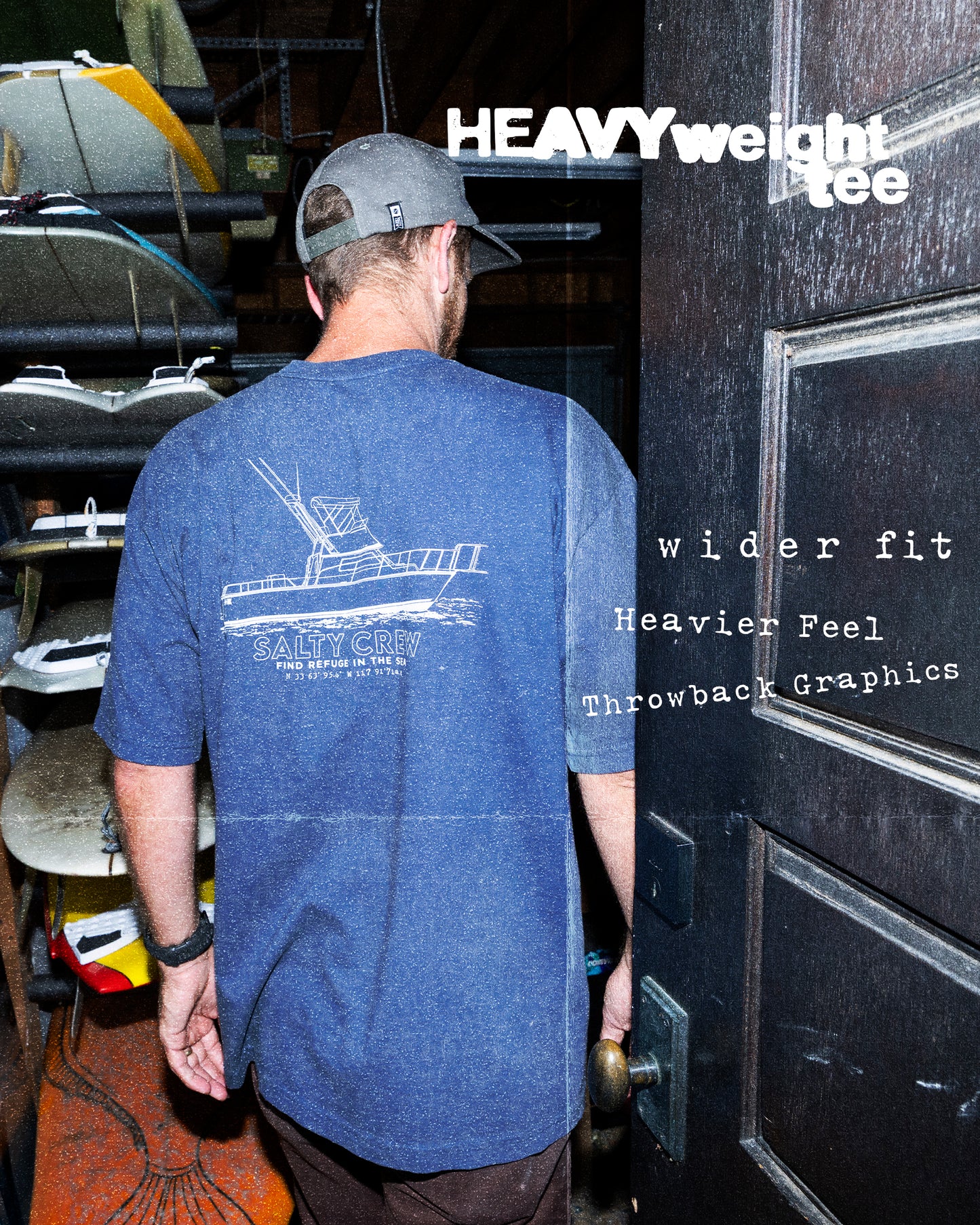 A man in a Washed Blue Vessel Heavyweight Tee and cap by Salty Crew stands by surfboards. The 100% cotton tee features a wider fit, heavyweight feel, and retro graphics.