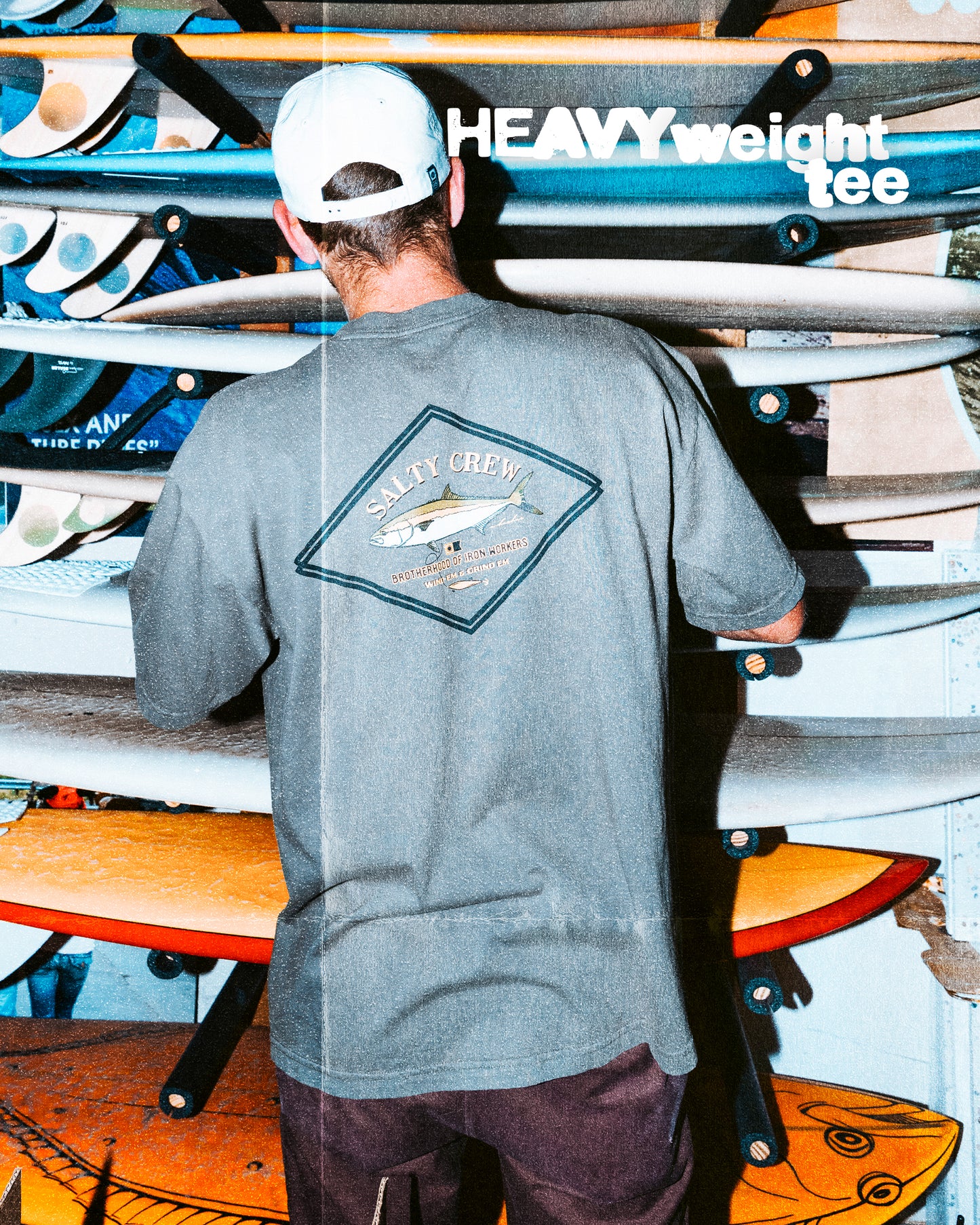 Wearing a gray Salty Crew Yellowtail Heavyweight Tee - Washed Dusty Olive and a white cap, someone stands by surfboards. Bold “HEAVYweight tee” text highlights the 100% cotton, screenprinted style for a laid-back, outdoorsy look.