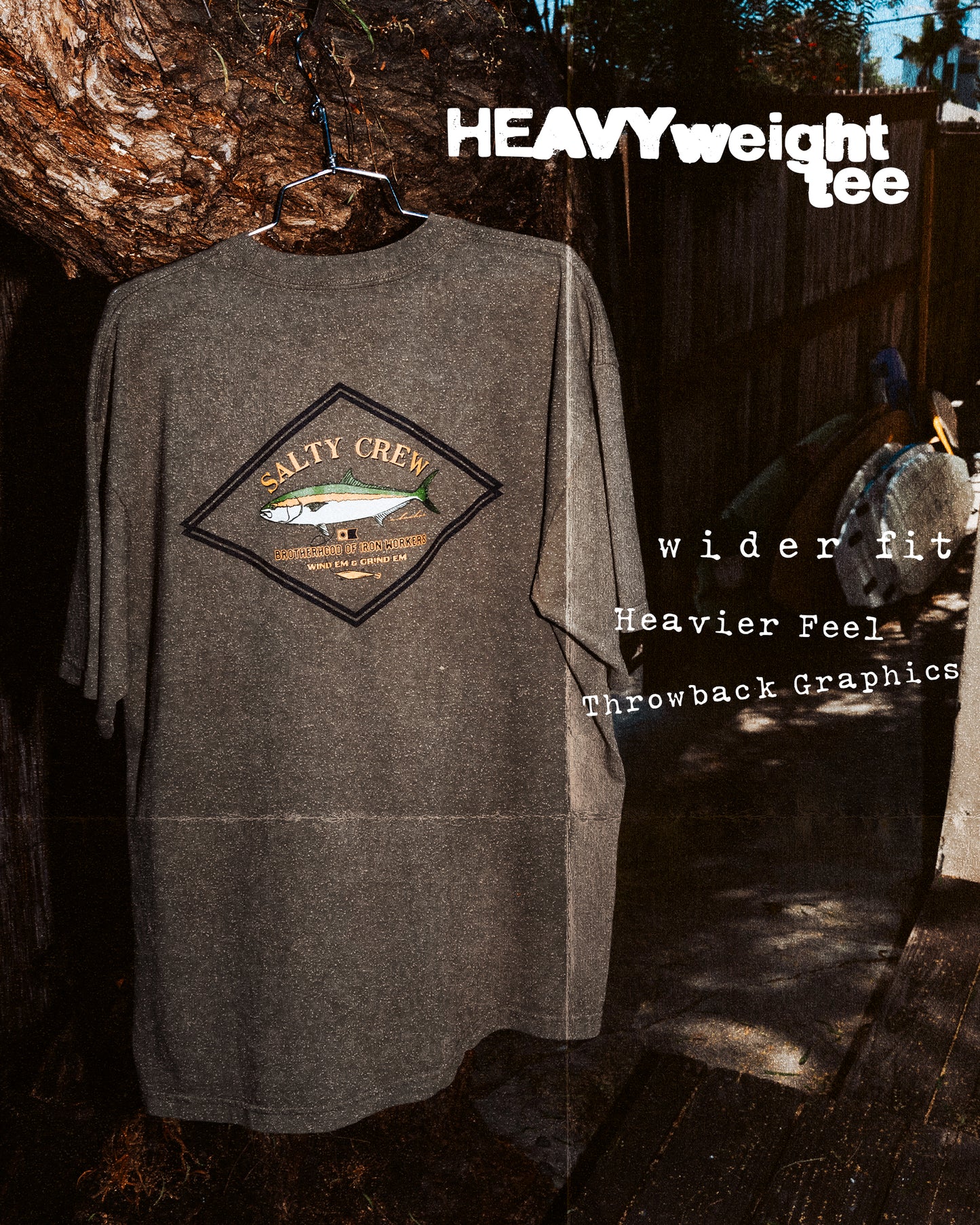 A Salty Crew Yellowtail Heavyweight Tee in washed dusty olive, 100% cotton with a fish graphic and throwback screenprinted text, hangs on a wire hanger outdoors. Features: heavyweight fabric, wider fit, heavier feel.