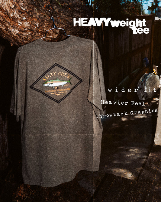 A Salty Crew Yellowtail Heavyweight Tee in washed dusty olive, 100% cotton with a fish graphic and throwback screenprinted text, hangs on a wire hanger outdoors. Features: heavyweight fabric, wider fit, heavier feel.