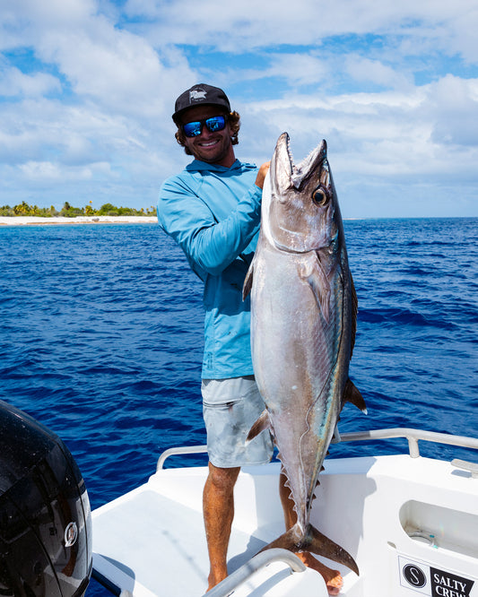 Wearing the Salty Crew Apex Perforated Sunshirt in Captains Blue, a person in sunglasses stands on a boat holding a big tuna, with blue ocean water and a tropical island in the background.