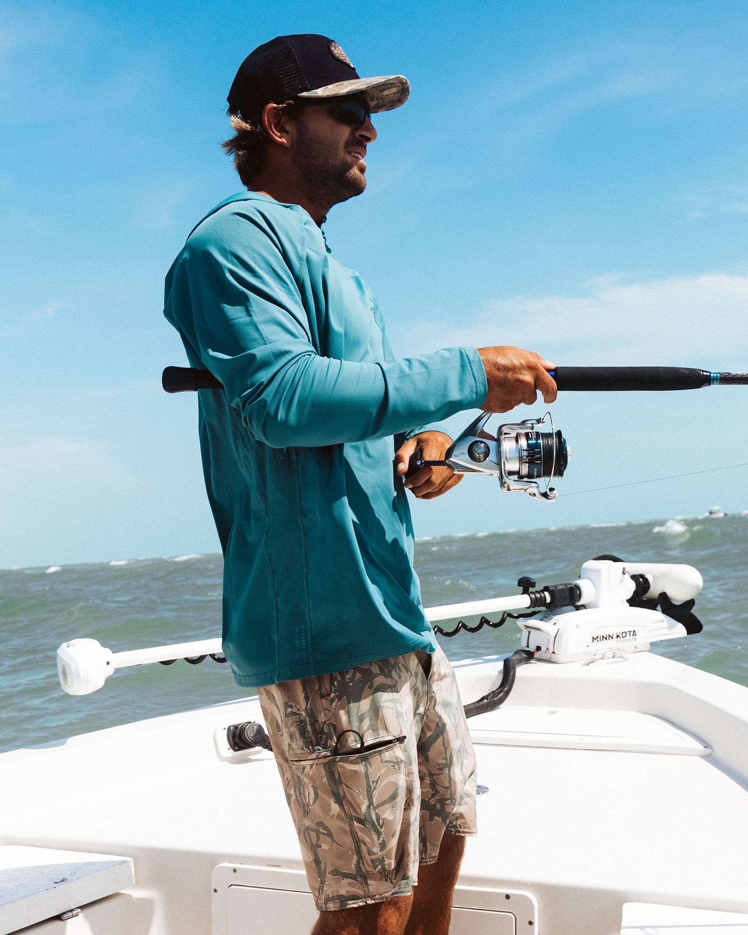 A man in a Salty Crew Apex Perforated Sunshirt - Hydro, camouflage shorts, and cap is fishing from a white boat on the ocean under a blue sky with scattered clouds.