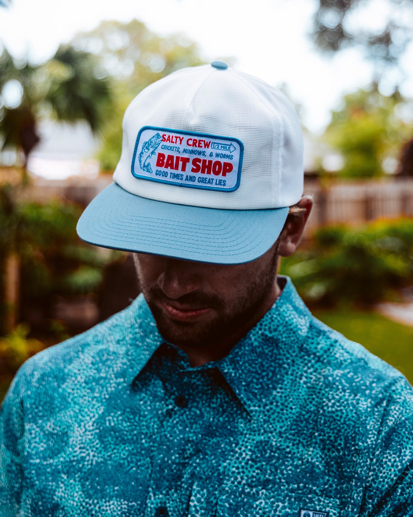 A man wearing the Salty Crew Bait Shop Trucker - Off White stands outdoors with his head slightly lowered, greenery and a wooden fence in the background.