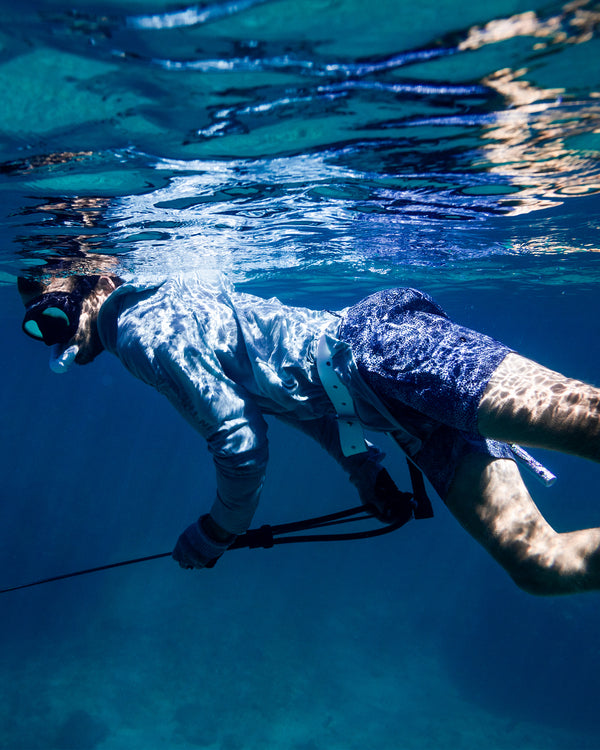 A person wearing a snorkel, mask, and rash guard swims underwater holding a speargun, surrounded by clear blue water with sunlight reflecting on their body.