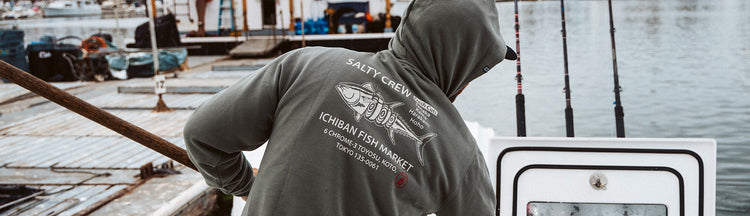 A person in a hoodie with a fish graphic and SALTY CREW text stands on a docked boat near fishing rods, with water and other boats in the background.