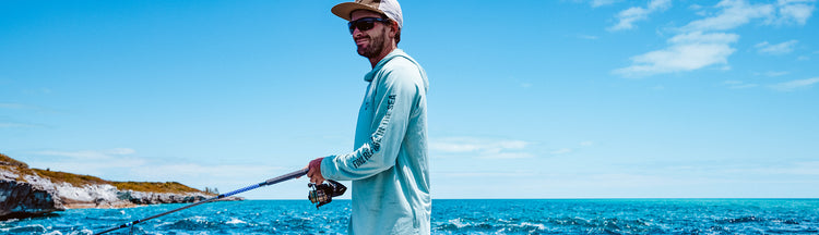 A man in a light blue long-sleeve shirt and hat smiles while fishing by the ocean, with rocky cliffs and a bright blue sky in the background.