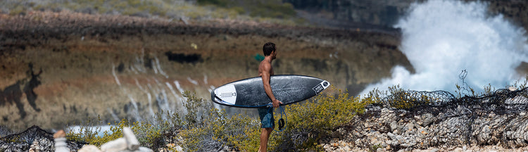 A man holding a surfboard stands on rocky terrain with sparse bushes, looking toward the ocean as a wave crashes in the distance.