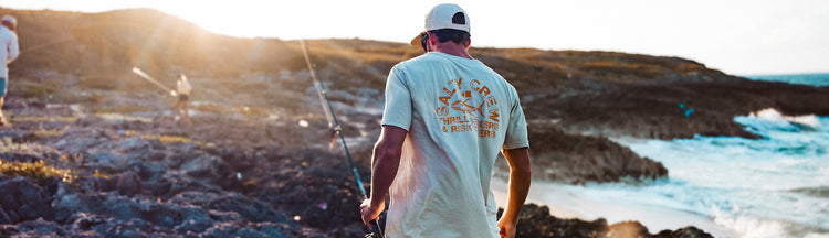 A person in a light t-shirt and white cap walks on rocky terrain near the ocean, holding a fishing pole. Another person is visible in the background. The sun is low, creating a warm glow.