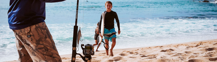 A person in camo shorts holds a fishing rod on a sandy beach, while another person in a wetsuit and blue swim trunks walks toward the shore carrying a surfboard. The ocean waves are in the background.
