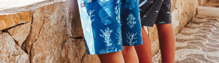 Two children standing side by side outdoors; one is wearing blue shorts with white coral patterns, while the other is wearing dark shorts. They are next to a stone wall, and part of a decorative stone path is visible.