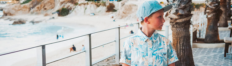 A young boy wearing a light blue cap and a patterned shirt stands near palm trees by a beach, looking to the side. The sandy shore and people relaxing by the sea are visible in the background.