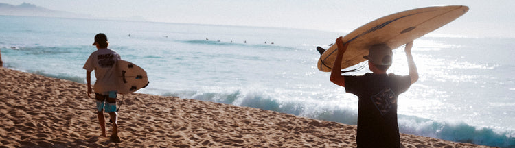 Two surfers walk on a sandy beach toward the ocean, each carrying a surfboard. The sun is shining, and gentle waves roll in, with distant mountains and surfers visible in the background.