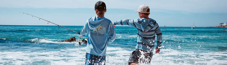 Two boys standing in shallow ocean water, one holding a fishing rod, both wearing hats and long-sleeve shirts, looking toward the sea on a bright, sunny day. Waves splash around their legs.