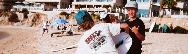 Two children wearing hats play together on a sandy beach near a resort, with surfboards and people relaxing under umbrellas in the background.