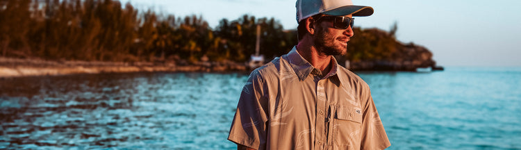 A man wearing sunglasses, a beige short-sleeve button-up shirt, and a white cap stands by the water at sunset, with trees and a boat in the background.