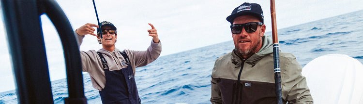 Two men are fishing on a boat in the ocean. One is smiling and gesturing excitedly while holding a fishing rod; both wear sunglasses, hats, and jackets. The sea and cloudy sky are visible in the background.