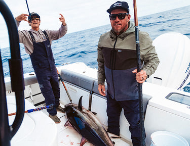 Two men on a boat at sea with fishing rods, standing next to a large tuna lying on the deck. One man is smiling and making hand signs; the other holds a pole and looks at the camera.