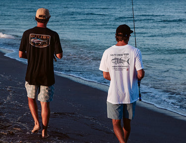 Two people in casual clothes and caps walk along the shore with fishing rods, facing the ocean. The water is calm, and the sun casts a warm light on the sandy beach. Both shirts have Salty Crew logos on the back.
