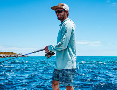 A man wearing sunglasses, a light blue long-sleeve shirt, and patterned shorts is fishing on a boat in bright, sunny weather with blue ocean waves and a distant shoreline in the background.