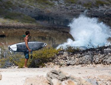 A man holding a surfboard stands on rocky terrain near the shore, gazing at large ocean waves crashing against the rocks in the background. Shrubs and fencing are visible around him.